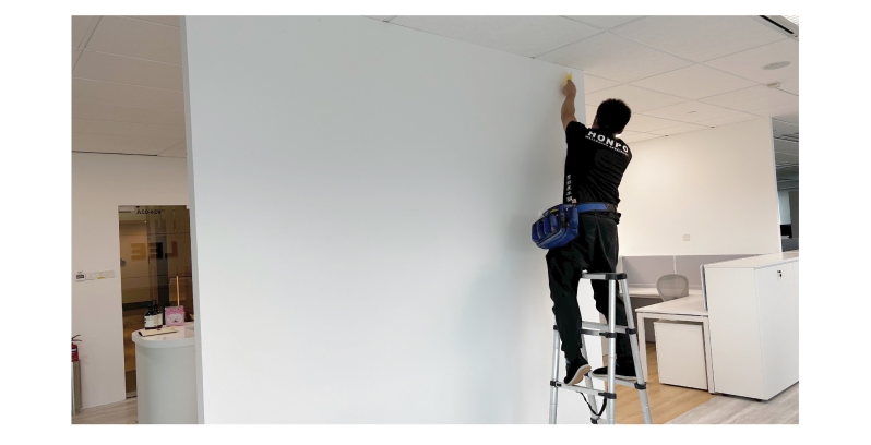 A person standing on a ladder while installing wallpaper on a white office wall